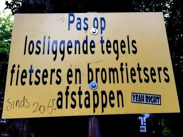 Waarschuwingsbord met de tekst: Pas op losliggende tegels fietsers en bromfietsers afstappen. Maar volgens een gestifte tekst dient men al sinds 2016 op te passen voor deze losliggende tegels. 